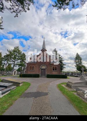 Cette vue plus large de la chapelle du cimetière de Malines englobe l'environnement sombre mais paisible où les pierres tombales et les monuments se dressent en compagnie tranquille. La chapelle, avec ses éléments gothiques et sa rosace centrale, préside la scène avec un air de révérence historique. Les chemins menant à la chapelle sont bordés de tombes, invitant à la réflexion sur les souvenirs et les legs qui y sont consacrés. L'image est un rappel poignant de l'histoire, du patrimoine, de la continuité de la vie et du souvenir. Veillée éternelle : la chapelle du cimetière de Malines. Photo de haute qualité Banque D'Images