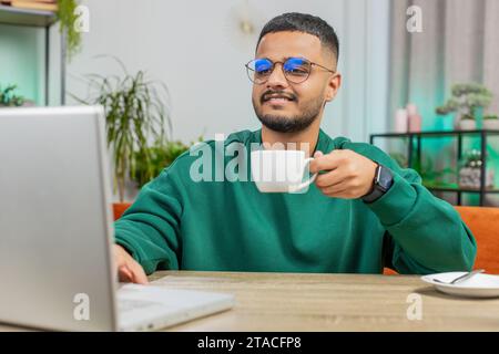 Homme indien souriant attrayant travaillant sur un ordinateur portable buvant une tasse de café chaud est assis à la table de travail à la maison salle de bureau dans la matinée. Homme hindou arabe appréciant se détendre, prendre une pause Banque D'Images