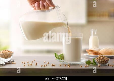 Remplissage du verre avec du lait de soja à partir d'une cruche sur la table de cuisine en bois avec des bols pleins de graines et une assiette avec du pain. Vue avant. Banque D'Images