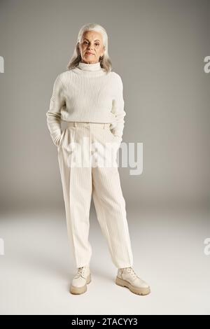 femme d'âge moyen aux cheveux attrayants et gris en tenue d'hiver blanche debout avec les mains dans les poches Banque D'Images