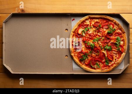 Pizza au pepperoni avec poivrons et roquette dans une boîte en carton ouverte sur une table de cuisine en bois naturel à plat avec espace de copie Banque D'Images