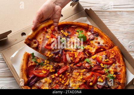 La main caucasienne humaine prend une tranche de grande pizza de la boîte de carton sur la table en bois blanc Banque D'Images