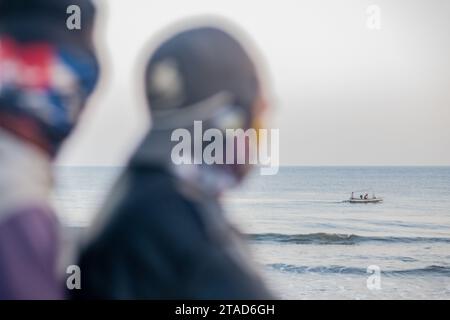 Deux personnages flous regardent vers la mer tandis qu'un bateau de pêche lointain navigue sur des eaux calmes sous un ciel doux et couvert Banque D'Images