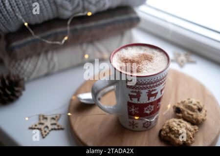 Nature morte de rebord de fenêtre d'hiver. Tasse en céramique rouge de café chaud sur le rebord de la fenêtre. Décorations de Noël sur le fond. Image de la maison confortable. Laine chaude kn Banque D'Images