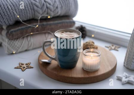 Nature morte de rebord de fenêtre d'hiver. Tasse en céramique bleue de café chaud sur le rebord de la fenêtre. Décorations de Noël sur le fond. Image de la maison confortable. Woolen k chaud Banque D'Images