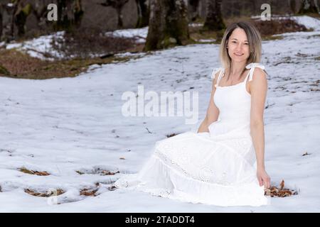 personne dans une robe blanche longue et fluide assise sur un rocher dans une forêt enneigée avec des arbres en arrière-plan Banque D'Images