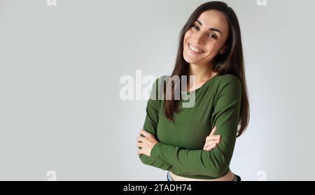 Portrait de jeune femme hispanique avec sourire toothy et bras croisés isolé sur mur gris avec espace copie. Magnifique fille en tenue décontractée regardant CAM Banque D'Images