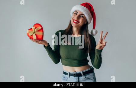 Heureux excité jeune femme magnifique dans le chapeau du père noël avec boîte-cadeau isolé sur fond gris. Bonne année 2024 et Noël. Shoppi en ligne Banque D'Images