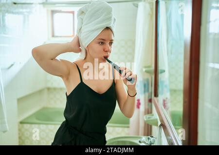 Jeune femme adulte dans la salle de bain avec une serviette sur la tête, favorise l'hygiène dentaire en utilisant une brosse à dents électrique, la réflexion miroir montre la routine de santé quotidienne. Banque D'Images