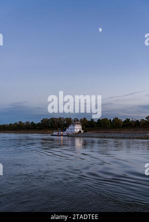 Barges de fret passant au-dessus des eaux calmes du fleuve Mississippi au crépuscule avec la lune haute dans le ciel Banque D'Images