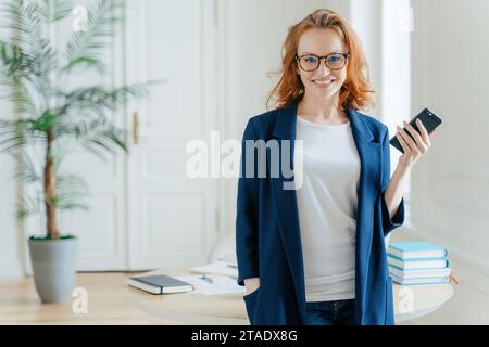 Femme aux cheveux rouges confiante en blazer bleu tenant un smartphone, à l'air professionnel Banque D'Images