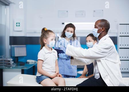 Médecin afro-américain portant un masque et une blouse de laboratoire vérifie la température d'une jeune fille. Le pédiatre masculin utilise un thermomètre et consulte la mère du patient. Banque D'Images