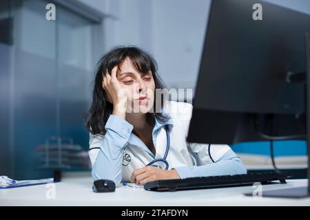 Jeune femme médecin portant une blouse de laboratoire et un stéthoscope, s'endormant devant un ordinateur dans le bureau de la clinique. L'air fatigué dans une chambre d'hôpital est un médecin caucasien avec la main sur son visage. Banque D'Images