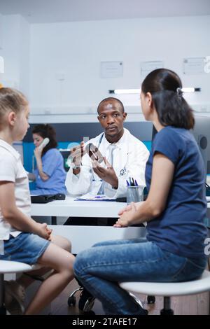 Tout en tenant une bouteille de médicament, un médecin afro-américain présente le traitement et le diagnostic à une femme caucasienne. Une mère et sa fille ont des consultations médicales. Banque D'Images