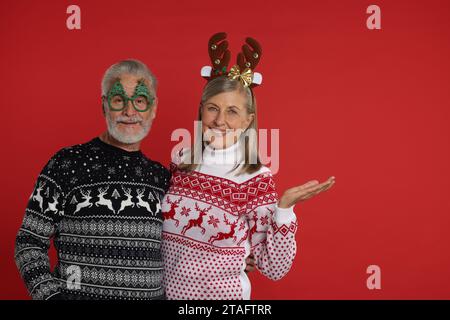 Couple senior en pulls de Noël, bandeau de renne et lunettes de fête sur fond rouge. Espace pour le texte Banque D'Images