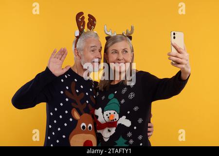 Couple senior en pulls de Noël et bandeaux de renne prenant selfie sur fond orange Banque D'Images