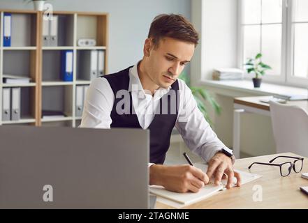 Jeune homme d'affaires sérieux travaillant dans le bureau et écrivant quelque chose dans un cahier Banque D'Images