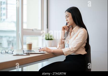 Une femme d'affaires asiatique spécialisée et professionnelle travaille sur son ordinateur portable au bureau, pensant et planifiant un nouveau projet. Banque D'Images