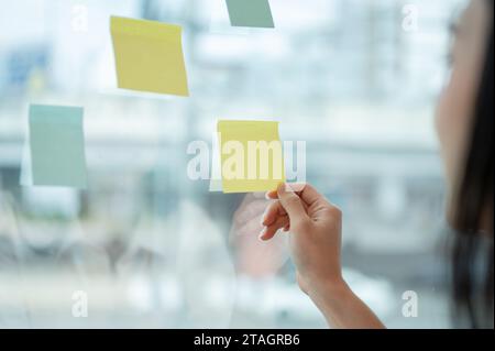 Image rapprochée d'une femme d'affaires ou d'une agente marketing attachant ou regardant une note collante sur un mur de verre. Une maquette de pense-bête vide. idée Banque D'Images