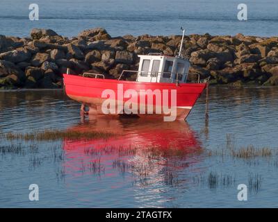 Pris à marée basse, un petit bateau rouge et blanc est ancré près des rochers. il bascule et se reflète dans les eaux peu profondes. Banque D'Images