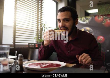 Concept de pizza Time. Employé de bureau africain masculin positif ou entrpreneur en costume mangeant un gros morceau de pizza Banque D'Images