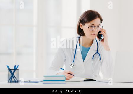 Médecin féminin concentré sur l'appel téléphonique avec ordinateur portable et notes dans le bureau Banque D'Images