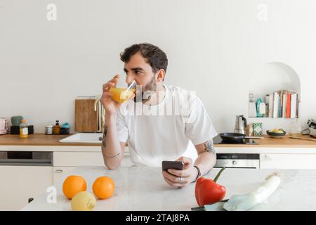 Homme barbu et tatoué en t-shirt blanc buvant du jus d'orange frais et utilisant un smartphone Banque D'Images