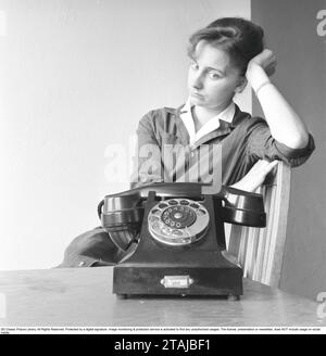 Femme dans les années 1950 Une jeune femme vue assise près du téléphone comme si elle attendait que quelqu'un appelle. À en juger par son expression faciale, elle commence à douter si elle sonnera avec un regard déçu sur son visage. Suède 1957 Banque D'Images