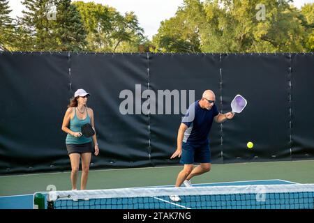 Un joueur de pickleball masculin renvoie une balle alors que sa partenaire féminine se tient près de la ligne de base. Banque D'Images