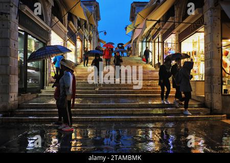 Les touristes sur le pont du Rialto avec des parapluies colorés passent devant les reflets illuminés du portique central, Venise sous la pluie, l'été Banque D'Images