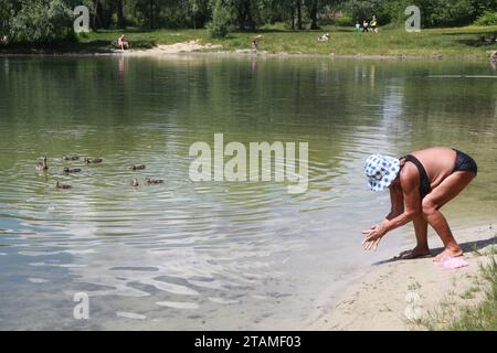 Femme caucasienne senior dans le chapeau de protection solaire se laver les mains dans Blue Lake à Kiev après avoir nourri une famille de canards près de la rive une belle journée en juin 2016 Banque D'Images