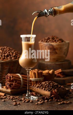 La crème irlandaise et la liqueur de café sont versées d'une bouteille dans un verre. Des grains de café, des bâtonnets de cannelle et de l'anis sont éparpillés sur la table. Banque D'Images