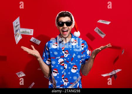 Photo de jeune homme d'affaires prospère porter chemise St nicholas imprimé célébrant Noël mouche billets de banque dollar isolé sur fond de couleur rouge Banque D'Images