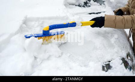 Gros plan d'une jeune femme brossant la neige de sa voiture un jour enneigé. Concept de l'hiver, de l'entretien de la voiture, et des tâches quotidiennes Banque D'Images