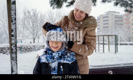 Jeune mère secouant la neige du chapeau de son fils après avoir joué dans le parc d'hiver. Amusement et joie pendant les vacances d'hiver, enfants jouant à l'extérieur, activité dans la neige Banque D'Images