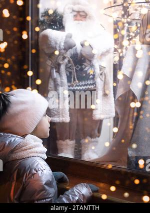 Fille regardant par la fenêtre aux décorations de Noël dans un magasin. Lumières de shopping de Noël en soirée Banque D'Images