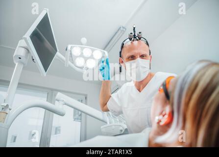 Rendez-vous en clinique dentaire dans un service médical moderne. Médecin dentiste dans des loupes pointant la lumière à la jeune femme prête pour la chirurgie des dents. Banque D'Images