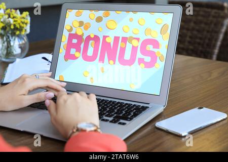 Gain de bonus. Femme utilisant un ordinateur portable à la table en bois à l'intérieur, closeup. Illustration de la chute de pièces de monnaie et de mot sur l'écran de l'appareil Banque D'Images