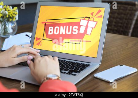 Gain de bonus. Femme utilisant un ordinateur portable à la table en bois à l'intérieur, closeup. Illustration de la boîte cadeau et mot sur l'écran de l'appareil Banque D'Images