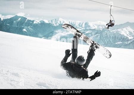 Snowboarder tombe sur le dos douloureusement avec des éclaboussures de neige sur la piste de ski hors-piste enneigée et vieux télésiège en arrière-plan. Journée ensoleillée d'hiver. Noir et Banque D'Images