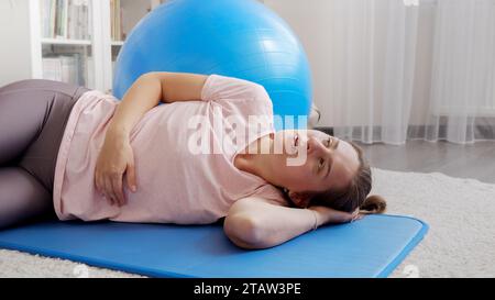 Femme en sueur fatiguée respirant à peine et ayant une pause avant de faire l'exercice de planche sur le tapis de fitness. Concept de soins de santé, sport et yoga à domicile. Banque D'Images