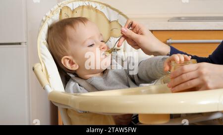 Gros plan du petit garçon devenant désordonné en mangeant du porridge dans une chaise haute. Concept de parentalité, nutrition saine et soins du bébé Banque D'Images