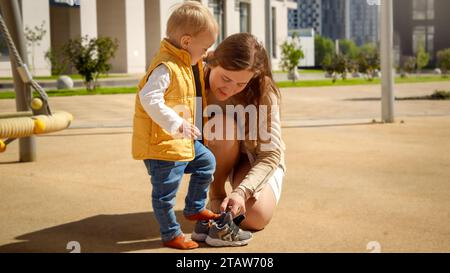 Jeune mère attentionnée aidant son tout-petit fils à mettre des chaussures sur le terrain de jeu après avoir joué à des jeux. Banque D'Images