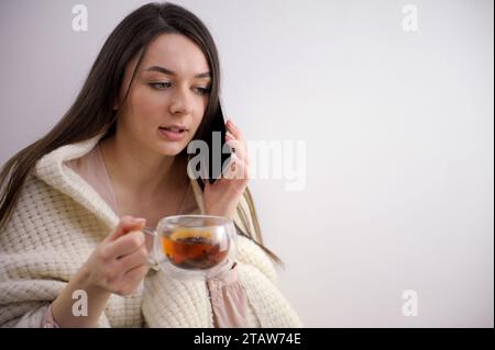 malade élégante femme au foyer de 20 ans en pull à col roulé et cardigan buvant une tasse de thé chaud avec gingembre, citron et miel sur fond bleu clair d'hiver. Photo de haute qualité Banque D'Images