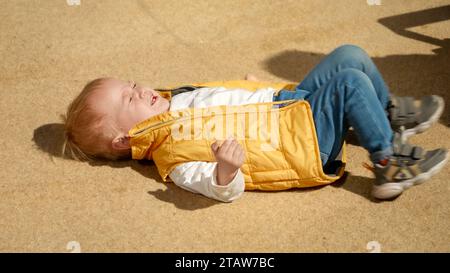 Pleurer et crier bébé garçon allongé sur le sol dans la rue. Enfants bouleversés, émotions négatives, problèmes d'enfants Banque D'Images