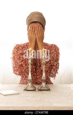 Femme juive prie sur des bougies de Shabbat allumées, couvrant son visage avec ses mains. A proximité se trouve un livre de prières religieuses. Banque D'Images