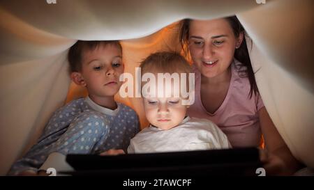 Mignon petit garçon avec la mère et le frère assis sous la couverture avec l'ordinateur tablette la nuit. Famille ayant du temps ensemble, parentalité, enfance heureuse et Banque D'Images