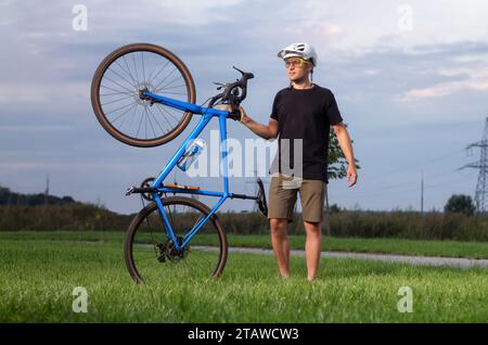 Heureux cycliste masculin portant casque et lunettes tient son vélo de gravier sur l'herbe verte dans le parc. Concept d'activités sportives. Banque D'Images