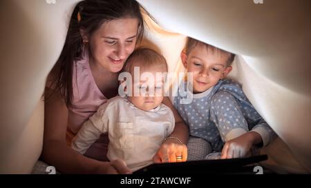 Deux garçons mignons avec la mère en pyjama jouant sur l'ordinateur tablette avec la mère sous la couverture dans le lit. Famille ayant du temps ensemble, parentalité, enfant heureux Banque D'Images