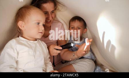 Deux garçons mignons se cachant sous la couverture avec la mère et jouant avec la torche. Famille ayant du temps ensemble, parentalité, enfance heureuse et divertissement Banque D'Images